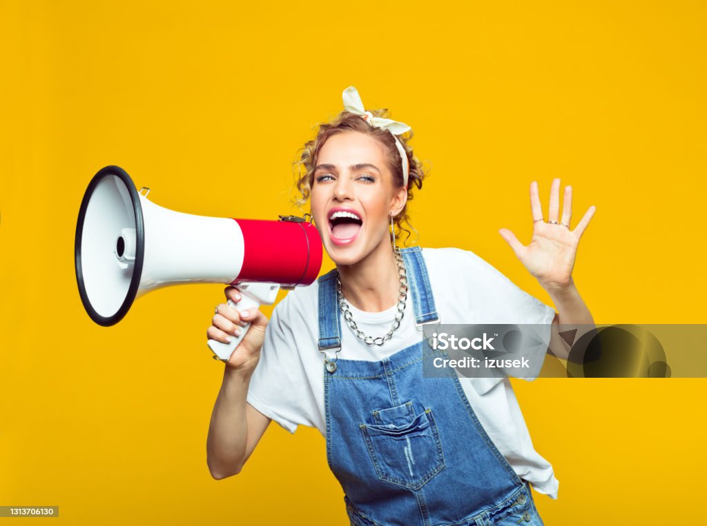 Young woman in dungarees shouting into megaphone Portrait of excited young woman wearing white t-shirt, denim dungarees and bandana shouting into megaphone. Studio shot on yellow background Women Stock Photo