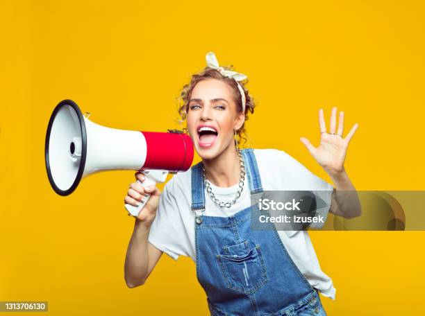 Young Woman In Dungarees Shouting Into Megaphone Stock Photo - Download Image Now - Women, Yellow, Megaphone