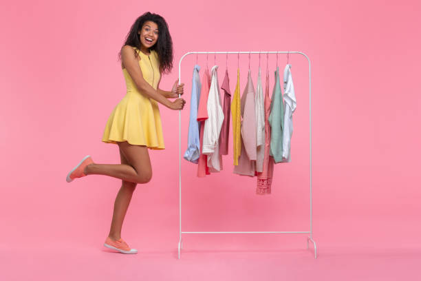 Full length portrait of cute happy smiling euphoric dark skinned girl wearing illuminating yellow mini dress choosing clothes wear hanging on white rack in showroom isolated on pastel pink background Full length portrait of cute happy smiling euphoric dark skinned girl wearing illuminating yellow mini dress choosing clothes wear hanging on white rack in showroom isolated on pastel pink background. dark pink dress stock pictures, royalty-free photos & images