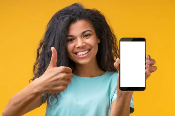 Photo of Cute african american girl with perfect smile holding mobile phone demonstrating blank white screen directly at the camera and holding thumb up.
