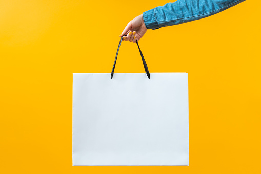 Cropped shot of woman hand holding plain white shopping bag on bright colored yellow background. Mock up, copy space for your text or logo.