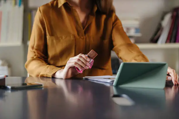 Photo of Unrecognizable Businesswoman Eating a Protein Bar at the Office, while Attending a Video Meeting with her Colleagues