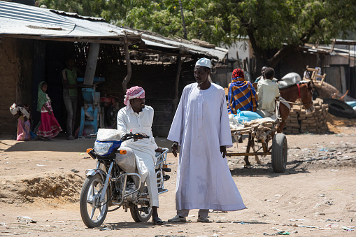 Bitkine, Chad - February 20, 2020: Two local men in traditional dress are standing in the main street of Bitkine talking, one of the men is on a motorcycle. Bitkine is an important trading place in Central Chad.
