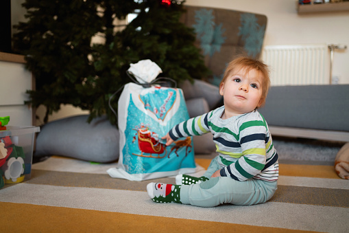 Cute Caucasian baby boy sitting on the floor with his present under the Christmas tree.