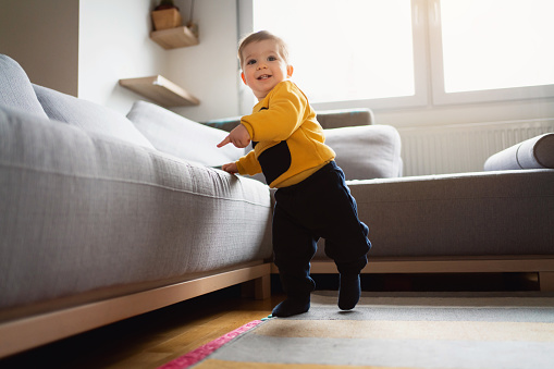 Low-angle view of a cute Caucasian baby boy making his independent steps at home while leaning on the sofa with his hand.