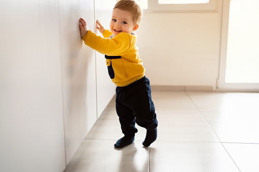 Cute Caucasian baby boy making his independent steps at home. He is walking on the tiled floor while leaning with his hands on the kitchen parts.