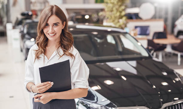 Female manager in car dealership Cheerful young woman with clipboard smiling and looking at camera while working in modern car dealership saleswoman stock pictures, royalty-free photos & images