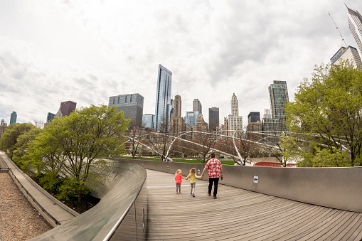 Chicago, IL, USA - April 18, 2021: A beautiful family walks along the BP Bridge, in Millennium Park, with the city skyline beyond.