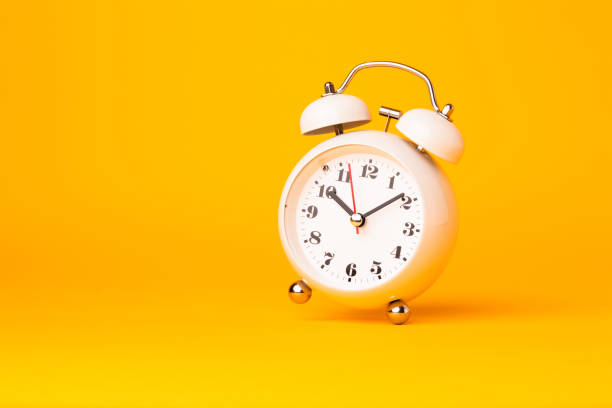 istock Small white alarm clock, black numbers, set the time placed on a table. Clock on isolated yellow background. 1313761353