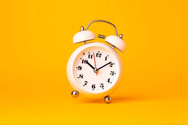 istock Small white alarm clock, black numbers, set the time placed on a table. Clock on isolated yellow background. 1313761364