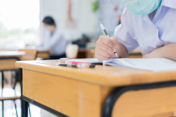 Asian Student girls wear face mask prevent COVID-19 coronavirus in Classroom for final test education exam room on High school. Students hold pencil writing paper documents for taking exams in room stock photo