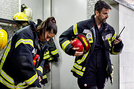 Hispanic woman and Caucasian man in 30s and 40s putting on turnout gear and using walkie-talkie as they prepare to depart for emergency.