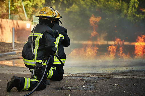 Full length rear view of first responders in turnout gear and helmets kneeling as they hold and support fire hose while putting out flames on asphalt.