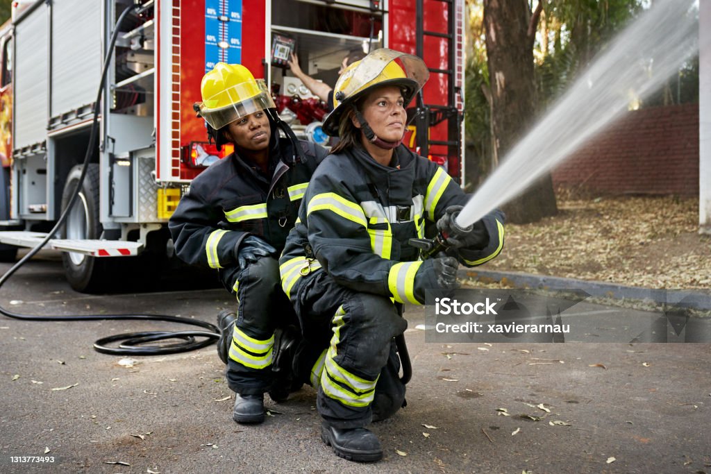 Mid Adult Female Hose Team Working at Emergency Site Full length view of Black firefighter providing backup for Hispanic teammate holding nozzle shooting straight stream of water. Firefighter Stock Photo