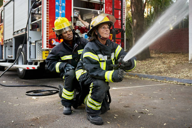 Mid Adult Female Hose Team Working at Emergency Site Full length view of Black firefighter providing backup for Hispanic teammate holding nozzle shooting straight stream of water. firefighter stock pictures, royalty-free photos & images