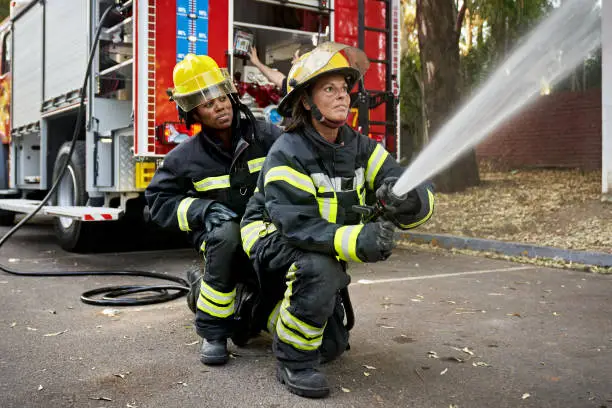 Full length view of Black firefighter providing backup for Hispanic teammate holding nozzle shooting straight stream of water.