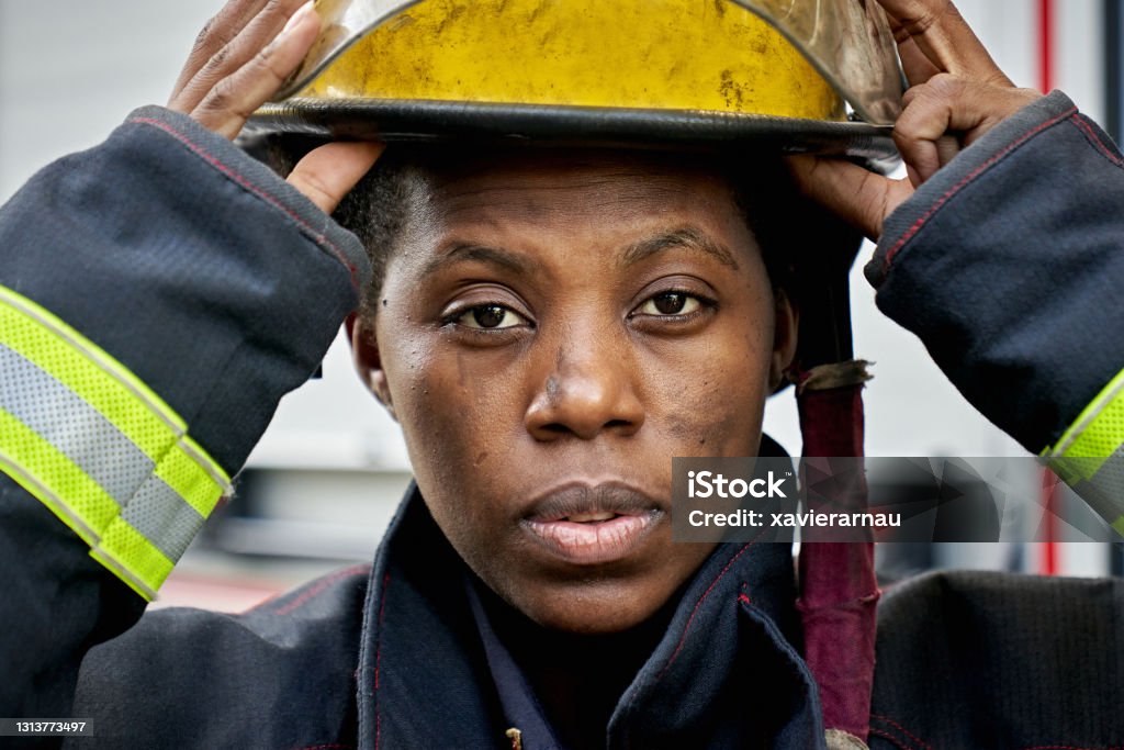 Portrait of Black Female Firefighter Putting on Helmet Close-up of mid adult first responder wearing protective workwear and looking at camera while securing helmet. Emergency Services Occupation Stock Photo