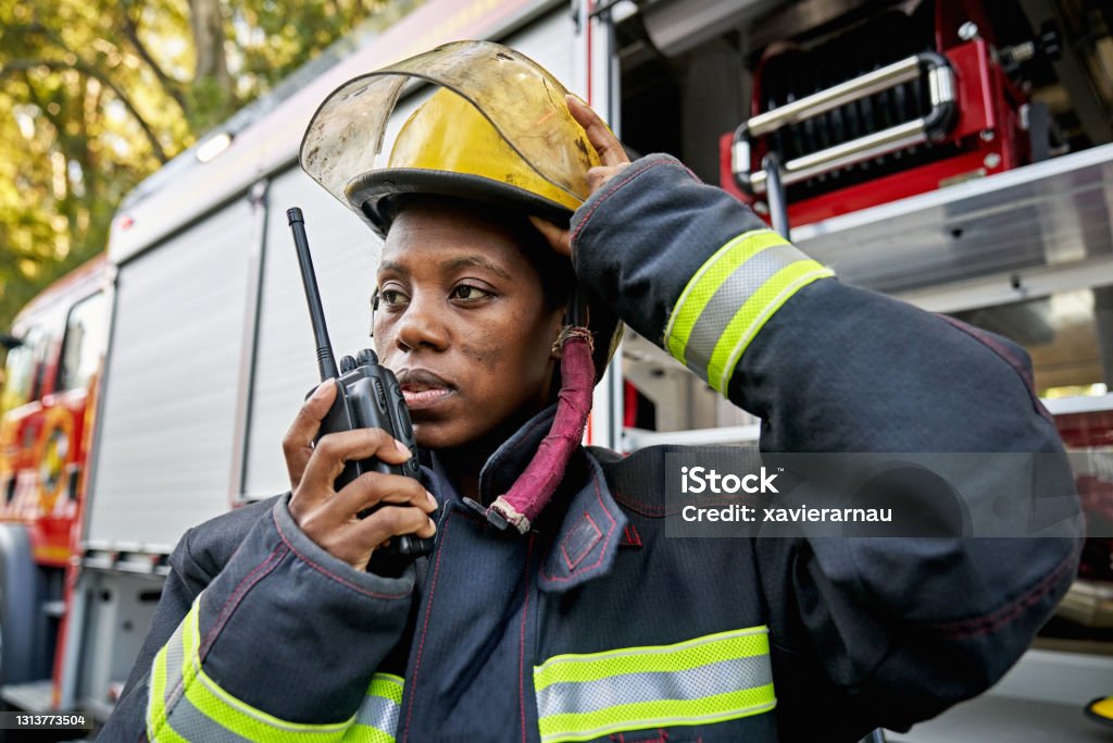 Black Female Firefighter Using Walkie-Talkie Waist-up view of first responder in early 30s wearing turnout gear, helmet, and standing next to fire engine while talking on portable two-way radio. Firefighter Stock Photo