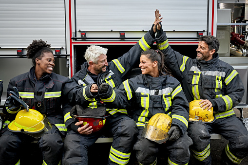 Front view of Black, Caucasian, and Hispanic first responders in 30s and 40s wearing turnout gear, holding helmets, and smiling as they support one another.