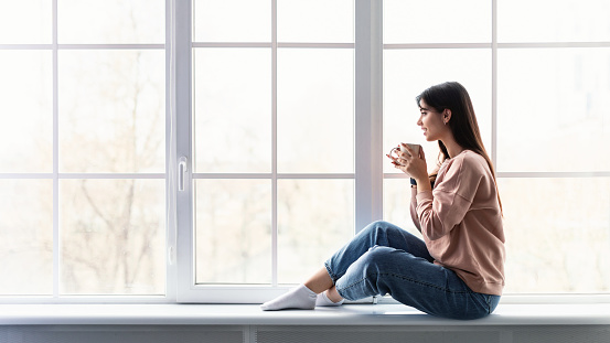 In The Morning. Profile of beautiful woman sitting on white windowsill at home, looking out of window and drinking hot coffee from cup. Happy lady taking break, holding mug with tea, copy space