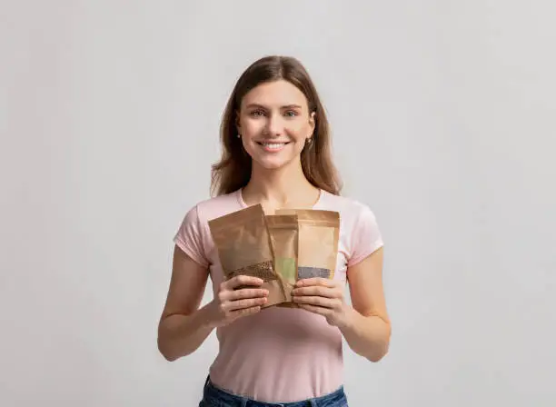 Eco Storage Concept. Smiling millennial lady holding set of brown kraft paper doypack bags with weight groceries, using biodegradable ziplock packs for food storing, standing over light background