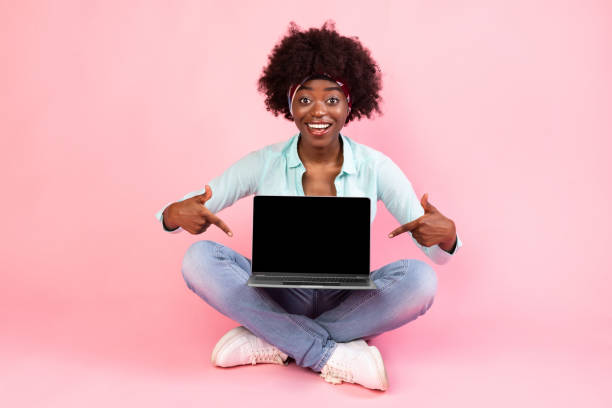 istock Black Young Woman Showing Laptop Empty Screen Sitting, Pink Background 1313787307