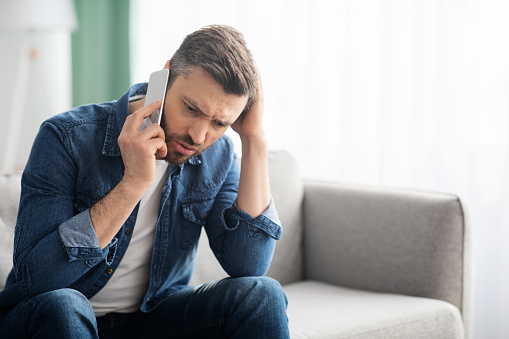 Worried middle-aged man having conversation on smartphone, touching his head, sitting on couch at home, panorama with copy space. Upset man calling his friends, asking for help
