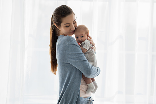 Beautiful young mother holding her adorable newborn baby while standing near window at home, loving millennial mom embracing her infant child, enjoying motherhood, copy space