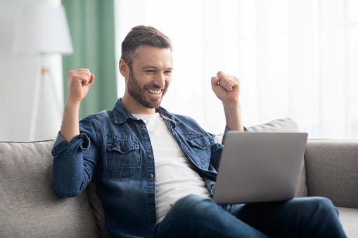 Emotional bearded middle-aged man celebrating success, gambling or trading online, using laptop while sitting on couch at home, copy space. Online gaming, financial markets concept