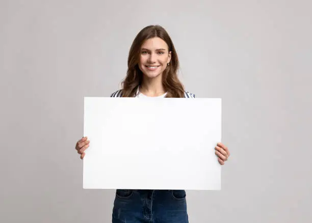 Smiling Millennial Lady Holding Blank Placard With Copy Space For Advertisement. Positive Young Woman Showing Empty Poster With Free Place For Your Text Or Design, Standing On Light Studio Background