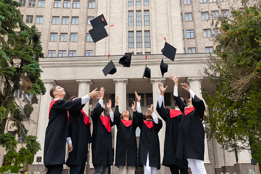 International group of students standing in a row and throwing graduation caps up in the air, posing next to university building after graduation ceremony, celebrating freedom