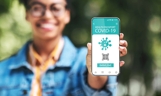 Ready For Travel. Smiling black young woman holding cellphone with digital health e-passport in hand, showing mobile application and confirming that she has immunity for covid-19, selective focus