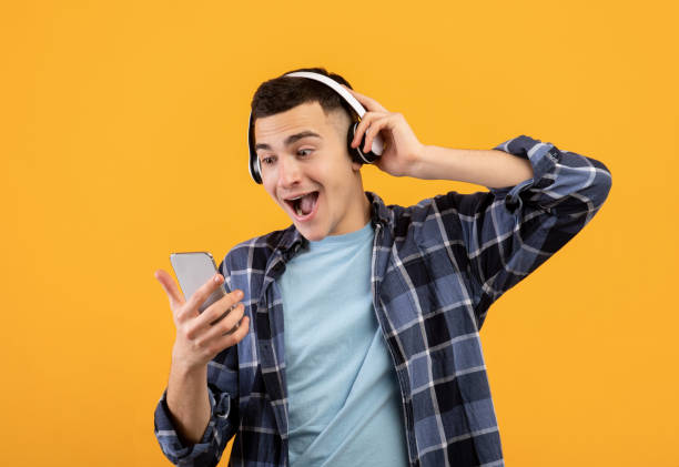 istock Excited Caucasian guy in wireless headphones using smartphone on orange studio background 1313796168