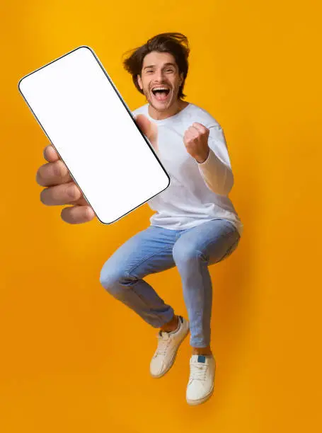 Photo of New awesome mobile app. Jumping emotional young guy showing smartphone with blank screen on orange background, mockup