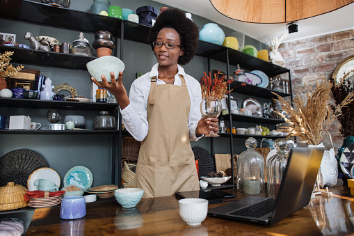 Beautiful afro american woman in eyeglasses and apron standing at store with various decor and using laptop for receipting of goods. Concept of people, technology and sale.