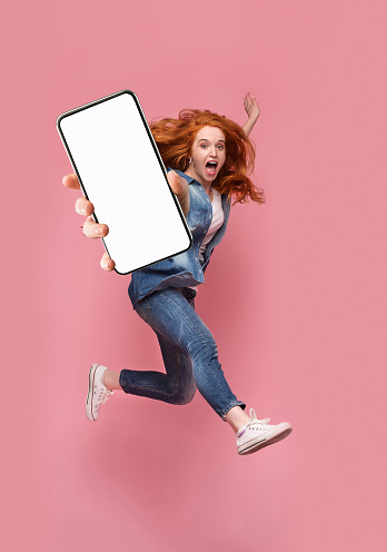 Excited redhead woman having fun, jumping,demonstrating mobile phone with empty white screen on pink studio background. Creative mockup collage with space for website or app design