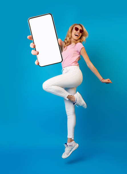 istock Pretty blonde lady holding smartphone, showing blank screen, jumping up over blue studio background, mockup for app 1313797537