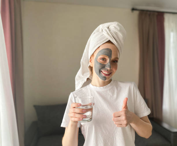 그녀의 머리에 수건과 그녀의 얼굴에 점토 마스크를 가진 예쁜 여자는 그녀의 다른 손에 물 한 잔을 들고 그녀의 손가락을 보여줍니다 - clear sky human skin towel spa treatment 뉴스 사진 이미지