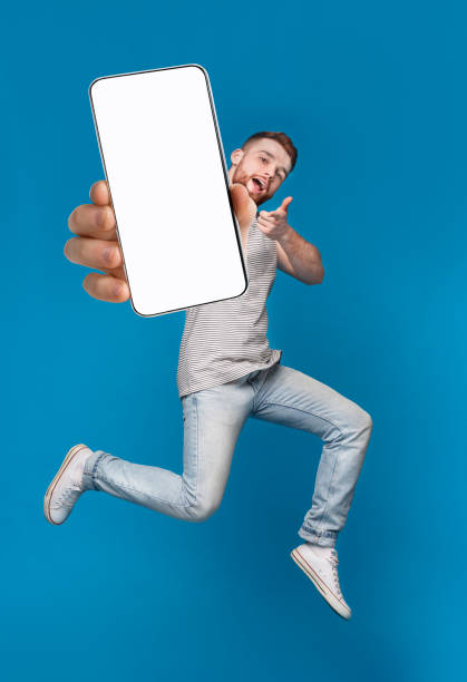 Emotional millennial redhaired guy pointing finger at camera, jumping with mobile phone on blue background, mockup