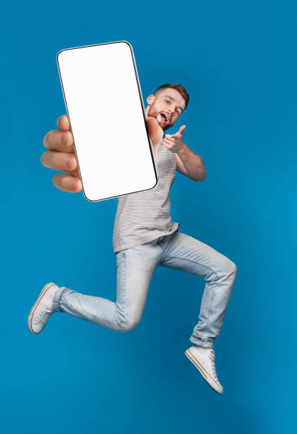 Emotional millennial redhaired guy pointing finger at camera, jumping with mobile phone on blue background, mockup Emotional millennial redhaired guy pointing finger at camera, jumping with mobile phone on blue studio background, mockup for mobile app on white screen. Creative collage with website template redhead photos stock pictures, royalty-free photos & images