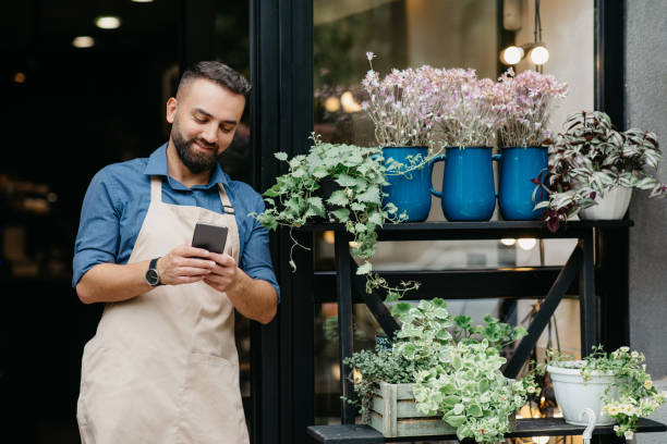 Portrait of young caucasian male owner of flower center typing on phone Portrait of young caucasian male owner of flower center typing on phone, employee manager in apron doing inventory. Cheerful man use smartphone near flower shop with plants in pots and vases outdoor florist stock pictures, royalty-free photos & images