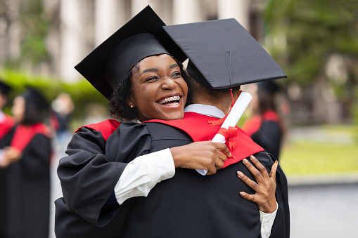Congratulations. Happy african american couple students in graduation dresses and hats embracing, having ceremony at university campus, enjoying and celebrating graduation, closeup portrait