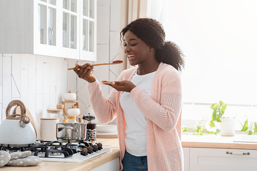 Cheerful Black Housewife Tasting Food While Cooking Healthy Lunch In Modern Kitchen, Happy African American Woman Enjoying Preparing Delicious Meal At Home, Smiling Lady Trying New Recipe, Free Space