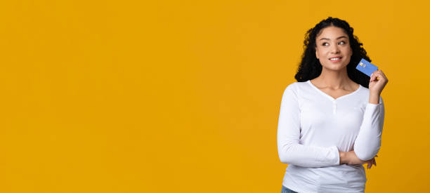 Ready For Shopping.Portrait of dreamy african american female holding credit card Ready For Shopping.Portrait of dreamy african american female holding credit card, pensive black woman planning purchases, standing isolated over yellow background, panorama with copy space thinking of you card stock pictures, royalty-free photos & images