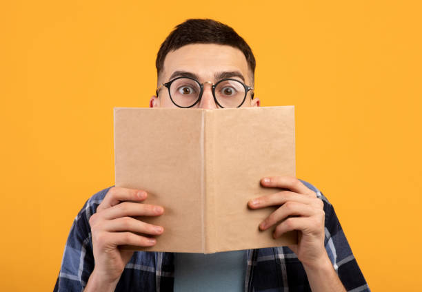 istock Shocked young guy in glasses hiding behind book, having problem studying for college exams on orange background 1313809278