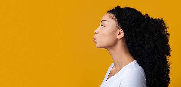 Profile Portrait Of Young African American Lady Sending Kiss, Side View Of Beautiful Black Woman Pouting Lips While Standing Isolated Over Yellow Background, Panorama With Copy Space