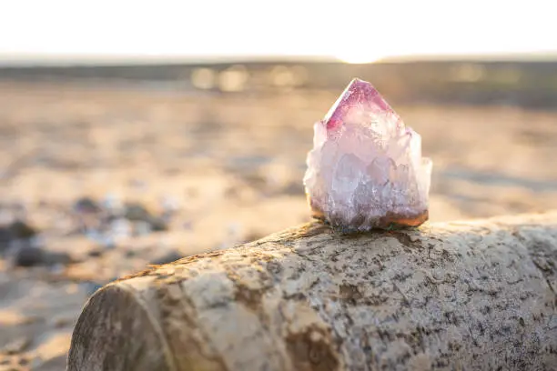 Amethyst crystal on wood, warm sunset beach background with copy space. Single raw natural purple geode outdoors, soft focus