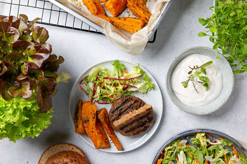 Grilled meat free plant based cutlets with sweet potato wedges, green mix salad and white sauce on gray table. Healthy vegan or vegetarian food concept. Top view.