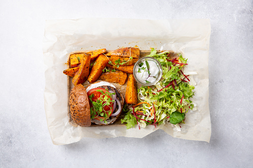 Meat free plant based burger served with sweet potato wedges, green mix salad and white sauce on gray table. Healthy vegan or vegetarian food concept. Top view.