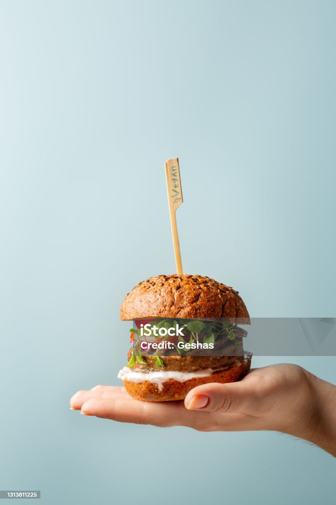 Hand holding burger. Hamburger with meat free plant based cutlet, tomatoes, onions, white sauce and microgreens on blue background. Healthy vegan or vegetarian food concept. Copy space. Burger Stock Photo